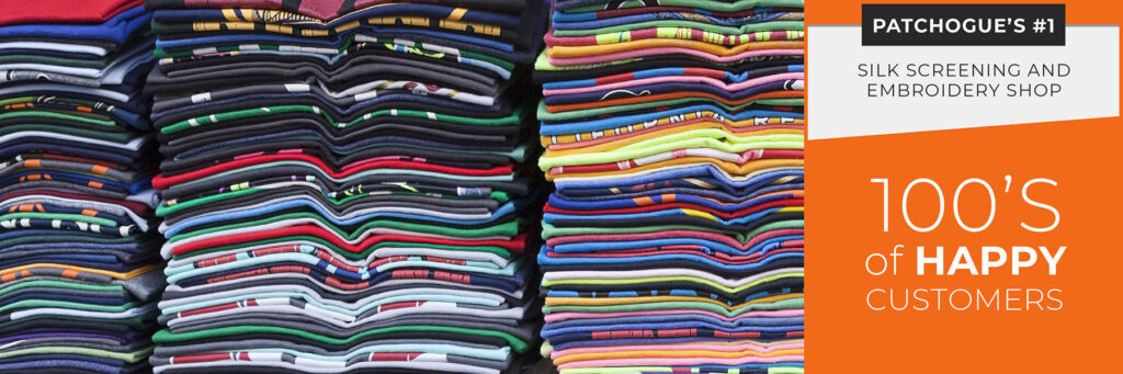 Stacks of folded, colorful custom t-shirts are shown next to a sign advertising a silk screening and embroidery shop known for expert screen printing and hundreds of happy customers in Patchogue.