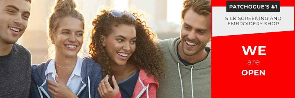 Four young adults smiling outdoors by a sign that reads: "Patchogue's #1 Silk Screening and Embroidery Shop. Custom t-shirts and embroidery, proudly serving Long Island. WE are OPEN.