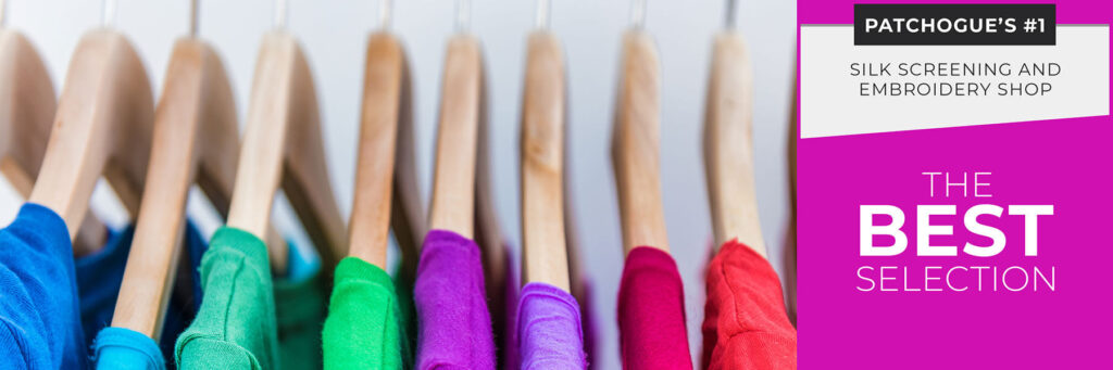 A row of colorful shirts on wooden hangers with a sign reading, "Patchogue's #1 Silk Screening and Embroidery Long Island Shop" and "The Best Selection of Custom t-shirts.