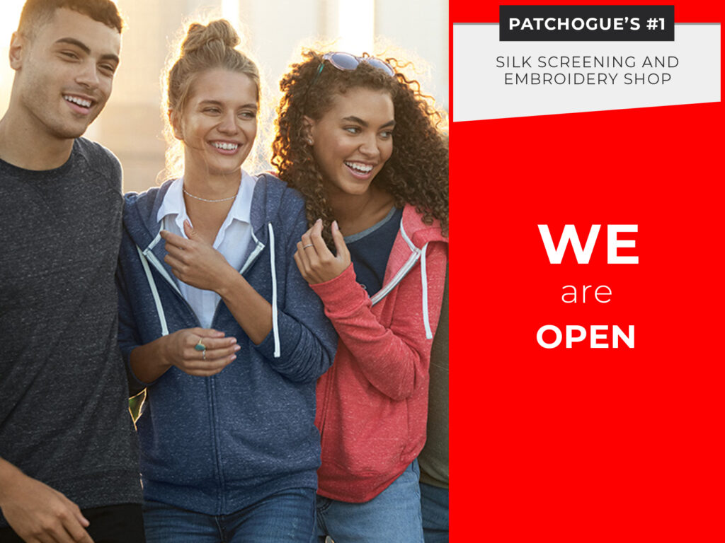 Three young adults in casual hoodies smile and stand together outdoors next to a sign reading, "Patchogue’s #1 Silk Screening and Embroidery Shop. We are open.”.