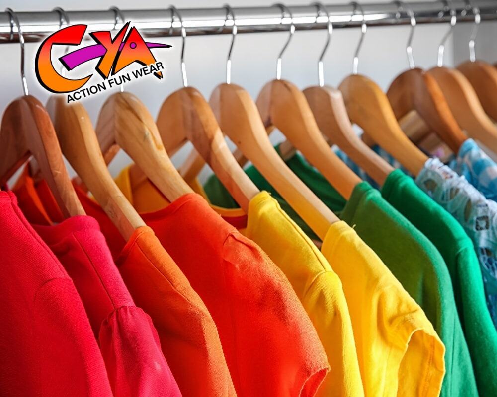 Colorful shirts sorted by shade hang on wooden hangers from a metal rack. In the top left corner, a “CYA Action Fun Wear” logo is displayed.