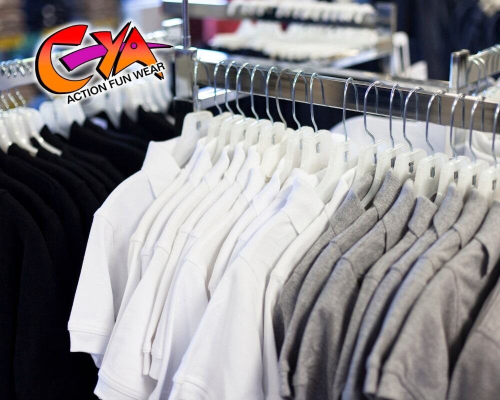 Rows of black, white, and gray t-shirts hang on racks, with a colorful "CYA Action Fun Wear" logo in the corner.